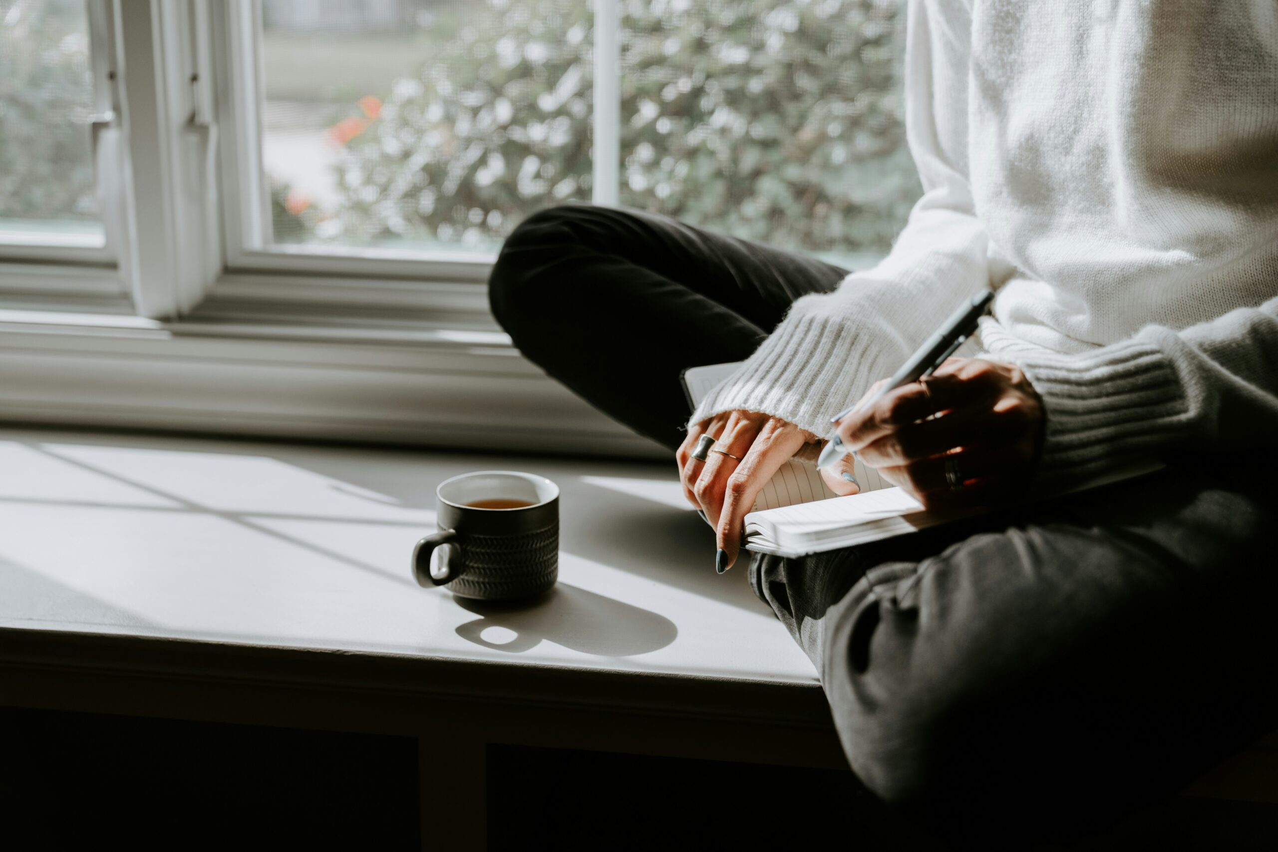 A serene photograph of a woman sitting peacefully by a window in soft natural light, gently holding her phone.<br />
