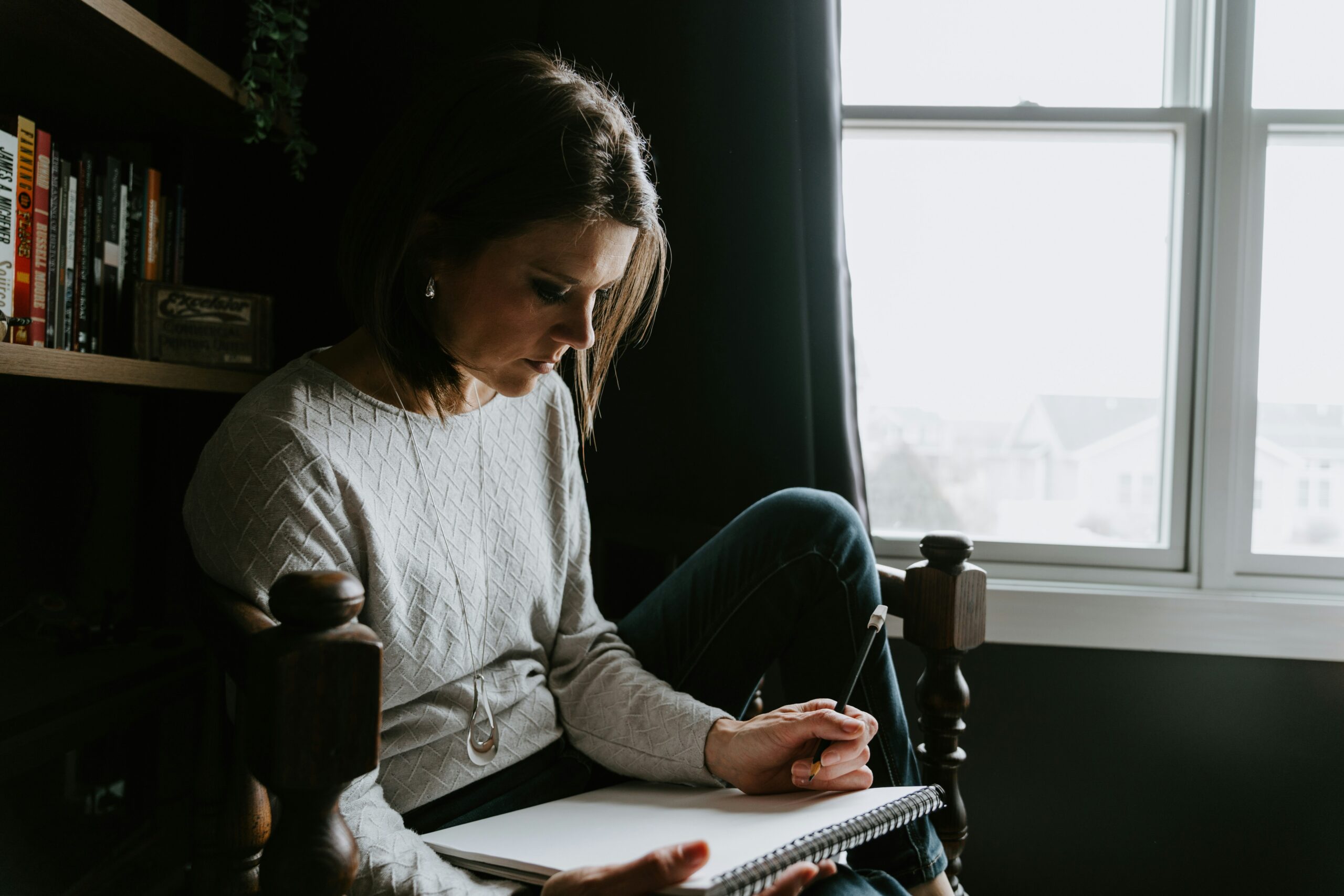 A calm scene of someone thoughtfully writing in a journal beside a window with soft morning light.<br />
