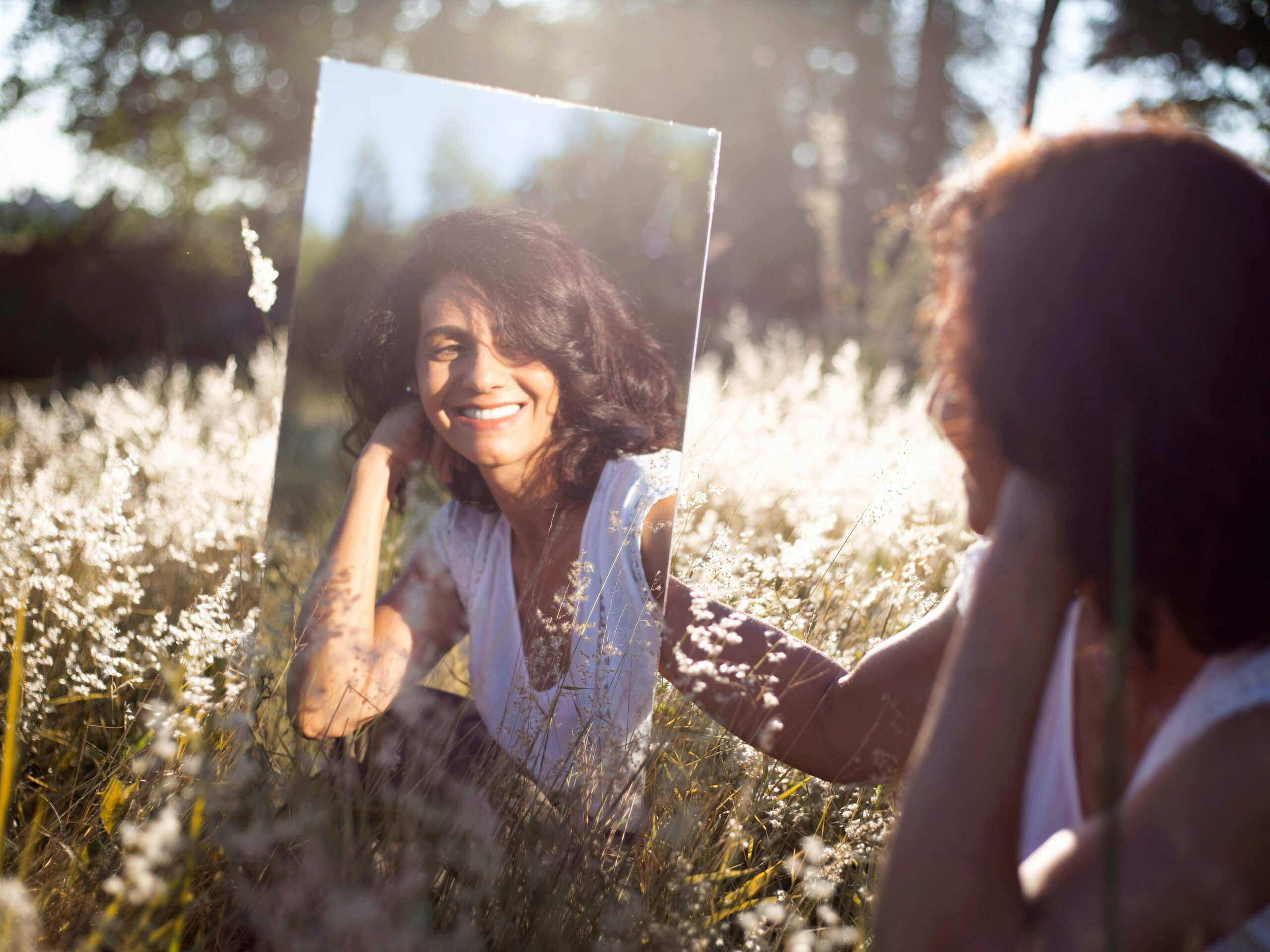 A warm photo of a woman smiling gently at her reflection in a mirror, bathed in soft natural light.<br />
